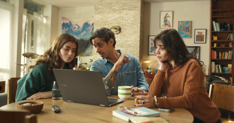 Tres personas sentadas en una mesa observando una notebook, en un ambiente hogareño cálido, representando el consumo de entretenimiento digital en familia