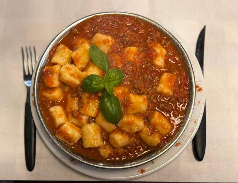 Plato hondo con ñoquis caseros en salsa bolognesa, coronado con hojas de albahaca fresca, servido en un bodegón típico con cubiertos al costado
