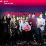 Grupo de personas celebrando con copas en mano sobre el escenario del VTEX Grand Prix 2025, con fondo iluminado y casco simbólico de premiación