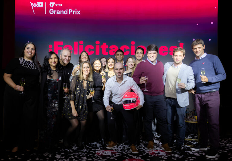 Grupo de personas celebrando con copas en mano sobre el escenario del VTEX Grand Prix 2025, con fondo iluminado y casco simbólico de premiación