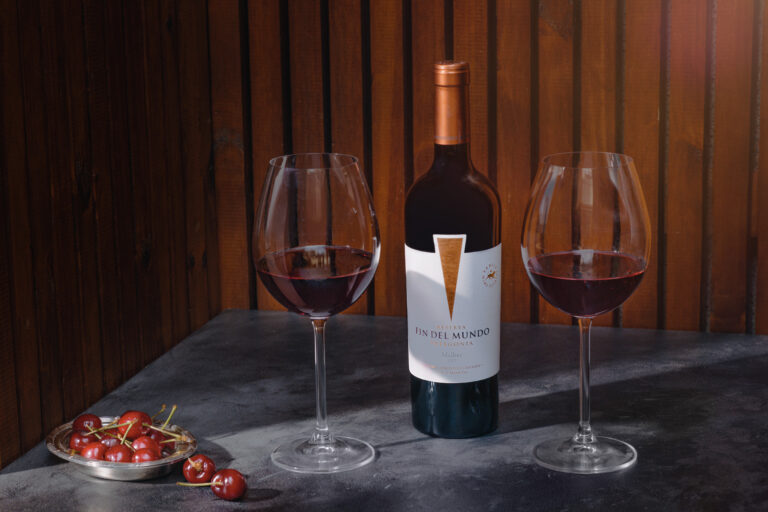 Botella de vino Malbec Reserva Del Fin Del Mundo acompañada por dos copas servidas y un plato con cerezas, sobre mesa oscura con fondo de madera, ideal para celebrar el Día del Padre con un vino patagónico