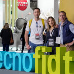 Asistentes posan junto al cartel de Tecno Fidta durante la feria de tecnología alimentaria, rodeados de stands y equipamiento industrial