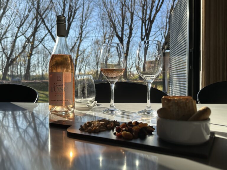 Botella de vino rosado Quinto Rosé Pinot Noir sobre mesa de madera, con copa servida y fondo rústico, representando la elegancia del vino argentino en mercados internacionales