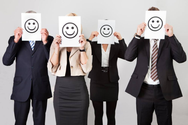 Grupo de empleados con trajes formales sosteniendo hojas con caras sonrientes frente a sus rostros, simbolizando el impacto positivo de los beneficios laborales en el clima organizacional
