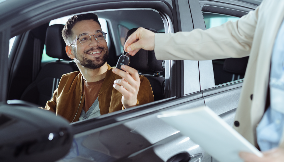 Un hombre sonriente recibe las llaves de su nuevo auto mientras permanece sentado en el vehículo, simbolizando una compra reciente y exitosa