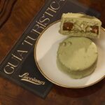 Primer plano de un alfajor verde sabor pistacchio de Lucciano’s sobre un plato blanco con borde dorado, apoyado en una mesa de madera junto a una guía turística