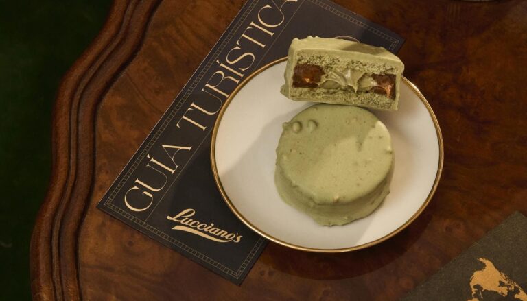 Primer plano de un alfajor verde sabor pistacchio de Lucciano’s sobre un plato blanco con borde dorado, apoyado en una mesa de madera junto a una guía turística