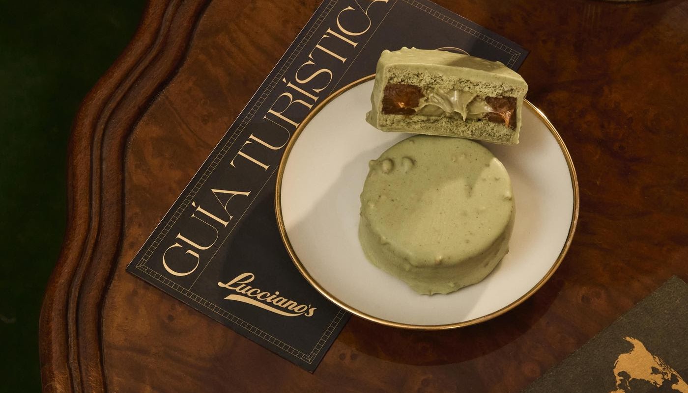 Primer plano de un alfajor verde sabor pistacchio de Lucciano’s sobre un plato blanco con borde dorado, apoyado en una mesa de madera junto a una guía turística