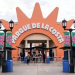 Vista frontal de la entrada del Parque de la Costa con su icónico cartel naranja y personas ingresando al parque