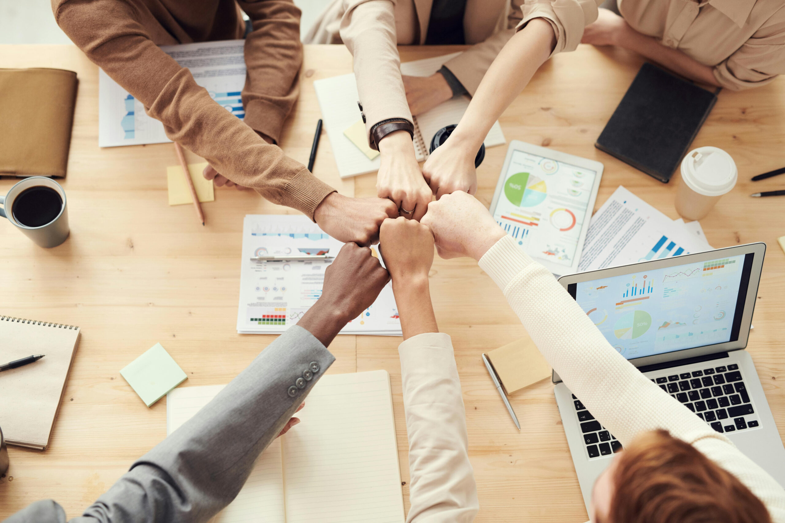 Manos de un equipo diverso unidas en un gesto de colaboración sobre una mesa con gráficos y computadoras, simbolizando el enfoque estratégico en Recursos Humanos.