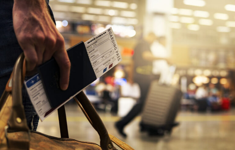 Hombre con bolso de mano sostiene pasaporte y tarjeta de embarque en un aeropuerto con personas y valijas desenfocadas al fondo