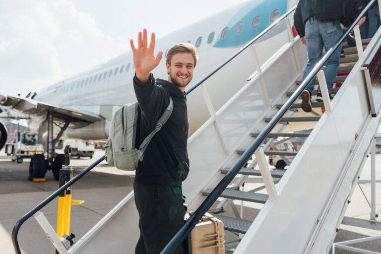 Joven viajero sonriente subiendo la escalerilla de un avión con mochila y valija, ilustrando la experiencia de viaje aéreo