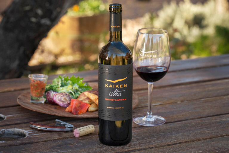 Botella y copa de vino Kaiken Ultra Cabernet Sauvignon sobre una mesa de madera, acompañadas de una tabla con vegetales y pan