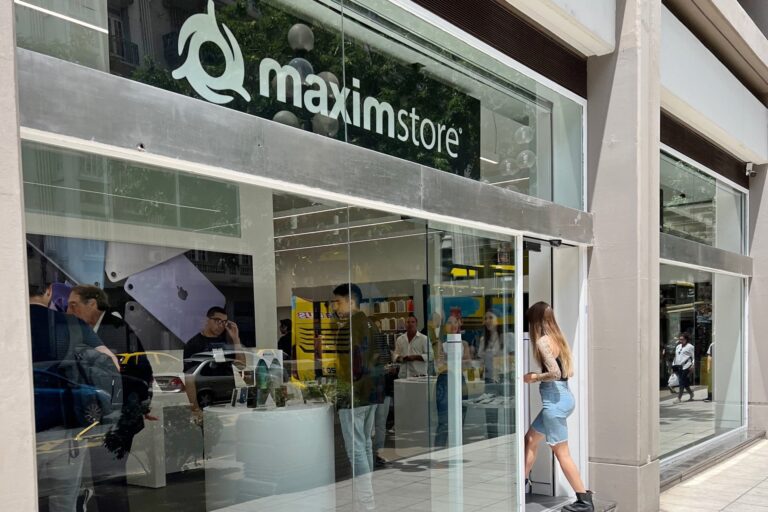 Fachada de la tienda Maxim Store con amplias vidrieras de vidrio, personas ingresando y productos tecnológicos en exhibición
