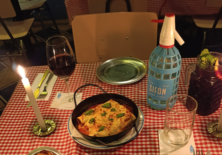 Mesa servida en Sifón con lasaña en cazuela, copa de vino, sifón de soda y bebida con menta sobre mantel rojo a cuadros, lista para una noche especial