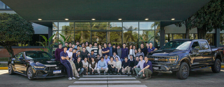Grupo de estudiantes posando en la entrada de un edificio junto a vehículos Ford Mustang y F-150 durante un workshop de innovación