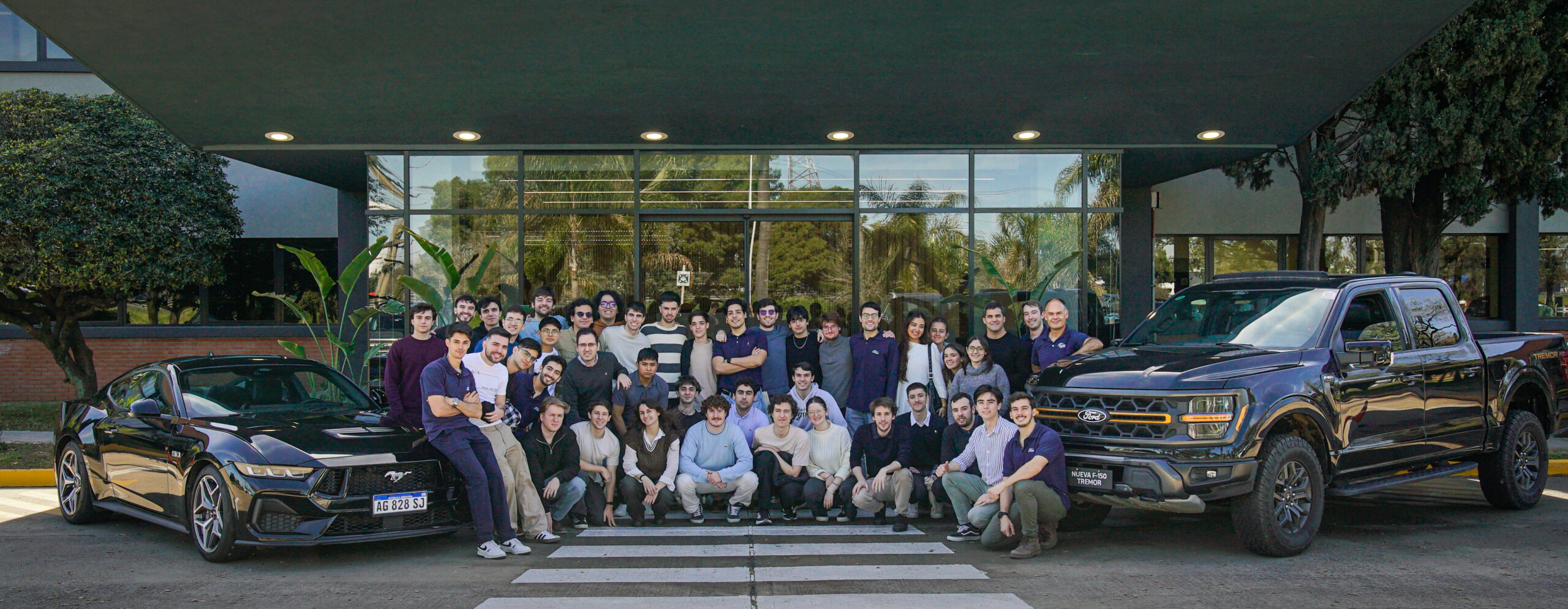 Grupo de estudiantes posando en la entrada de un edificio junto a vehículos Ford Mustang y F-150 durante un workshop de innovación