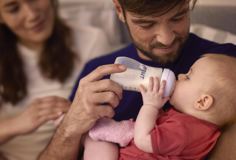 Padre alimentando a su bebé con un biberón, acompañado de la madre, representando el cuidado y bienestar familiar