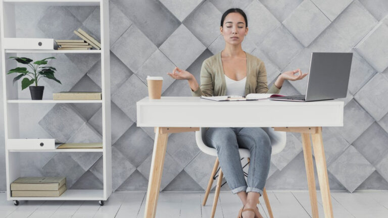 Mujer meditando en su escritorio con notebook, café y libros, representando el bienestar laboral en la oficina