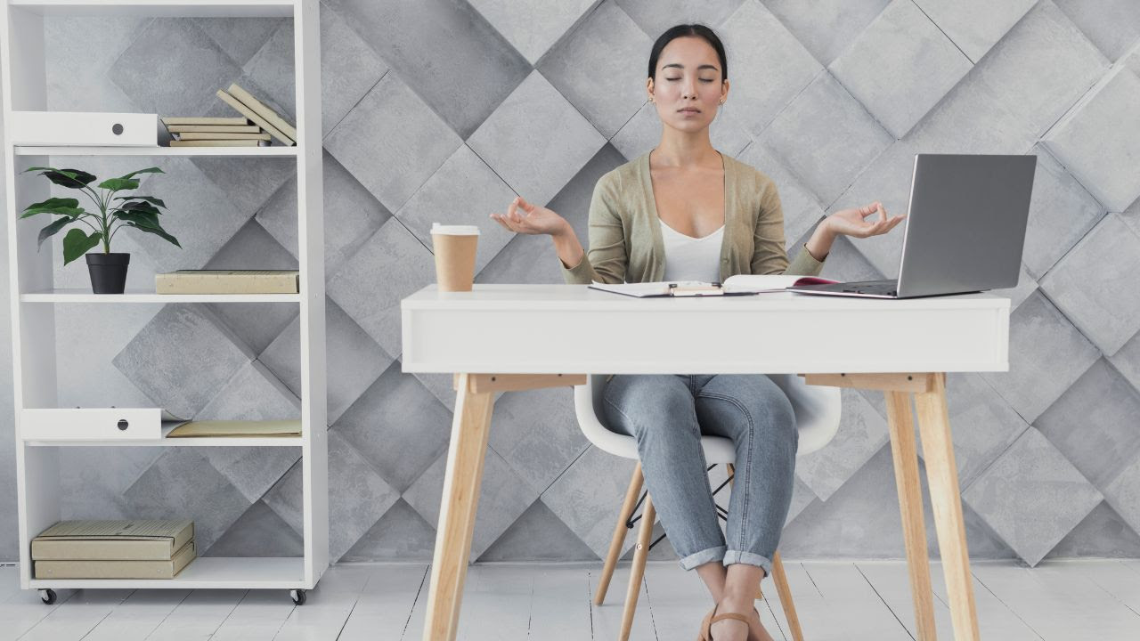 Mujer meditando en su escritorio con notebook, café y libros, representando el bienestar laboral en la oficina