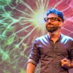 Hombre con micrófono de audífono dando una charla sobre un escenario, frente a una proyección colorida de fondo