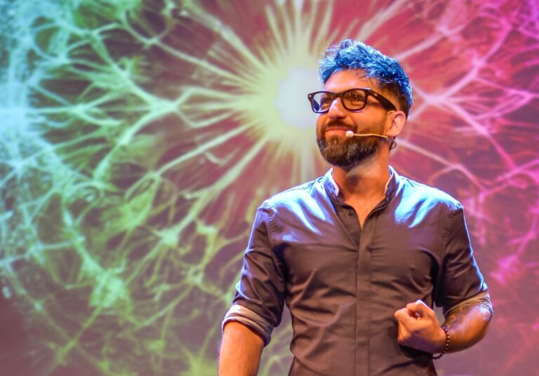 Hombre con micrófono de audífono dando una charla sobre un escenario, frente a una proyección colorida de fondo