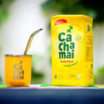 Mate de calabaza con bombilla junto a yerba en un cuenco y un paquete amarillo, sobre una mesa de madera en ambiente cálido