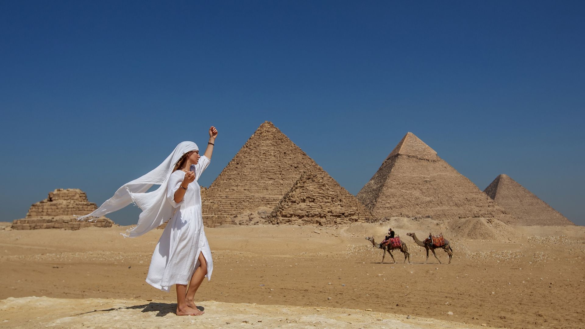 Mujer caminando frente a las pirámides de Egipto con camellos al fondo en un paisaje desértico