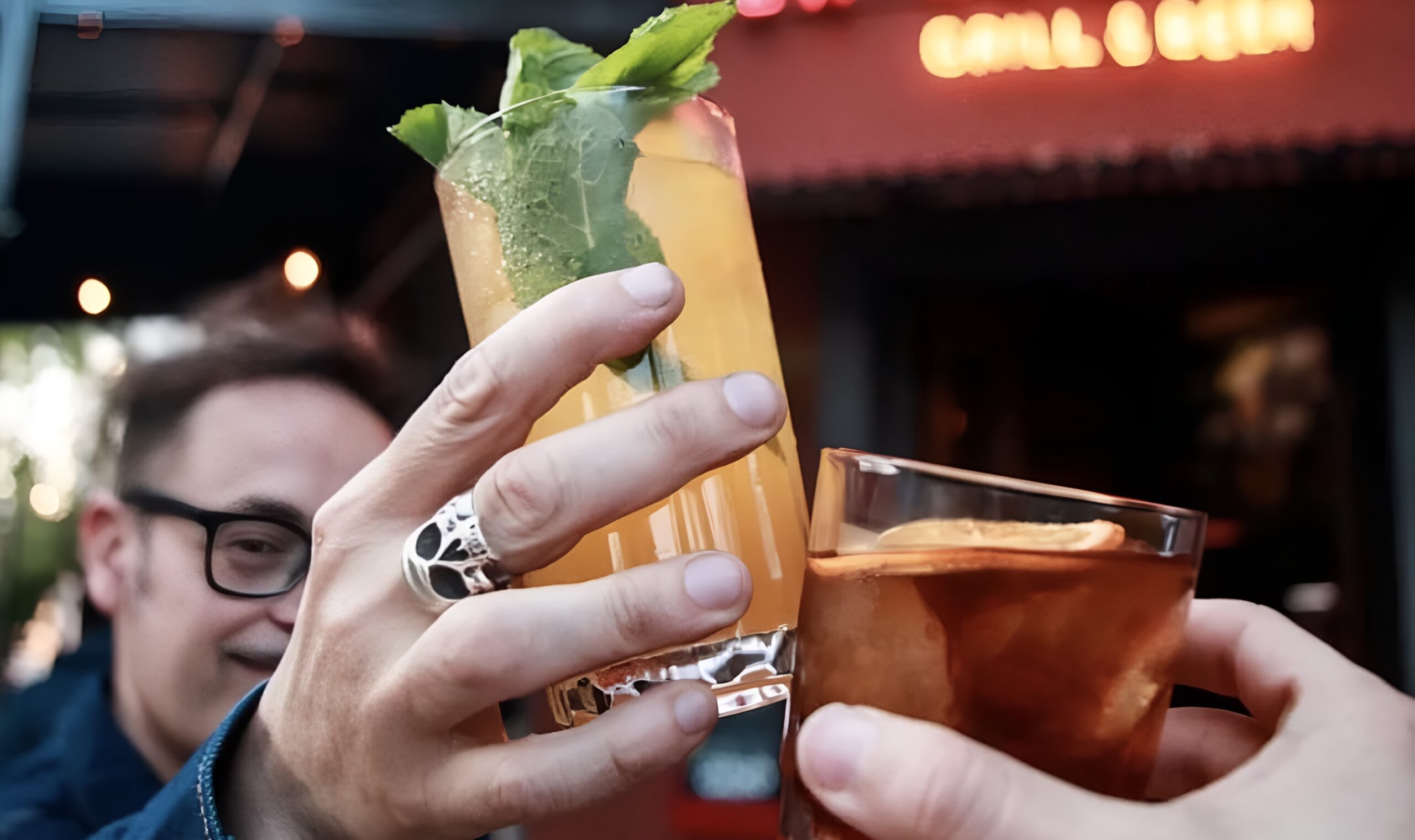 Dos personas chocando vasos con cocktails en primer plano, frente a un bar iluminado