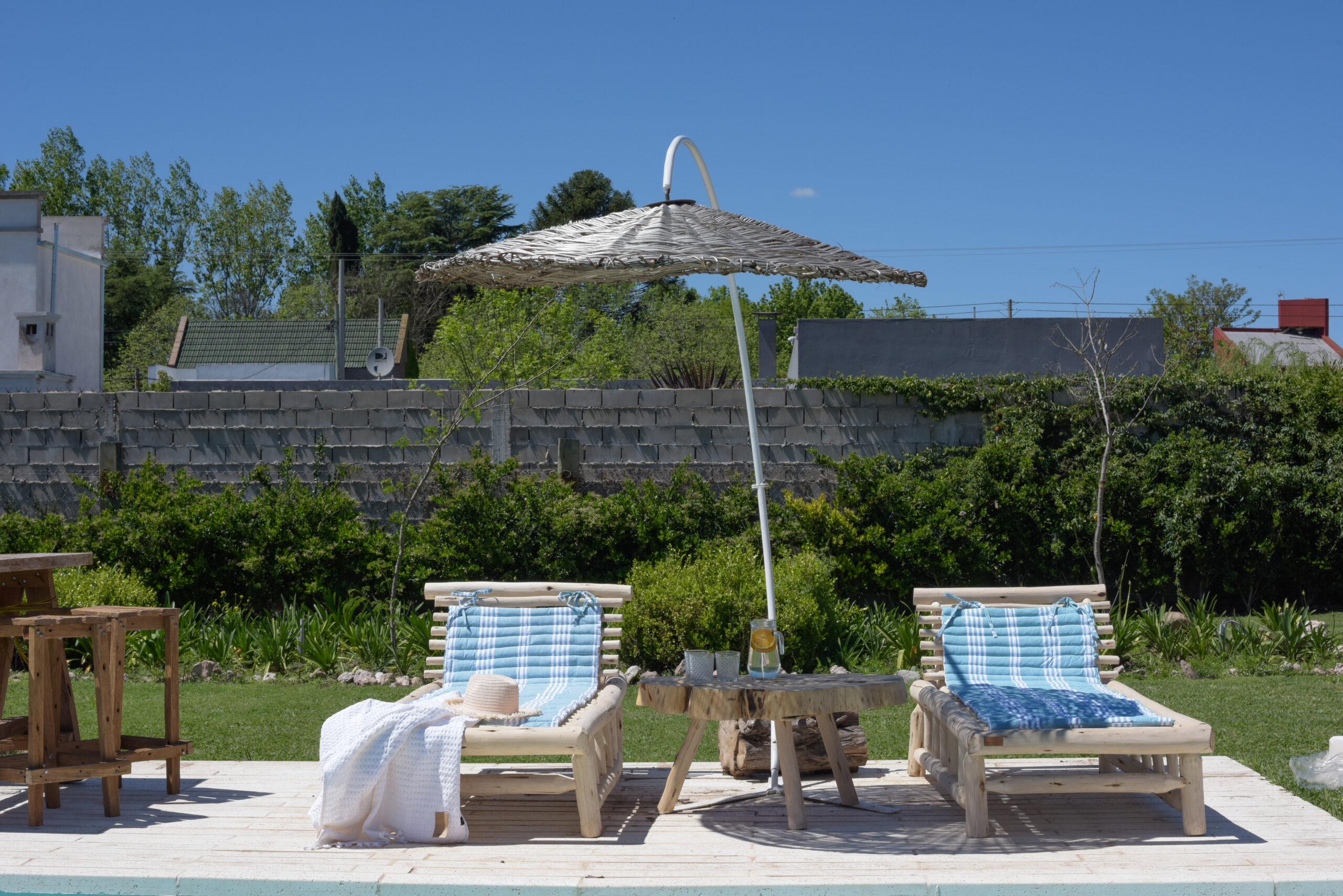 Reposeras de madera con almohadones y sombrilla junto a una piscina en un jardín