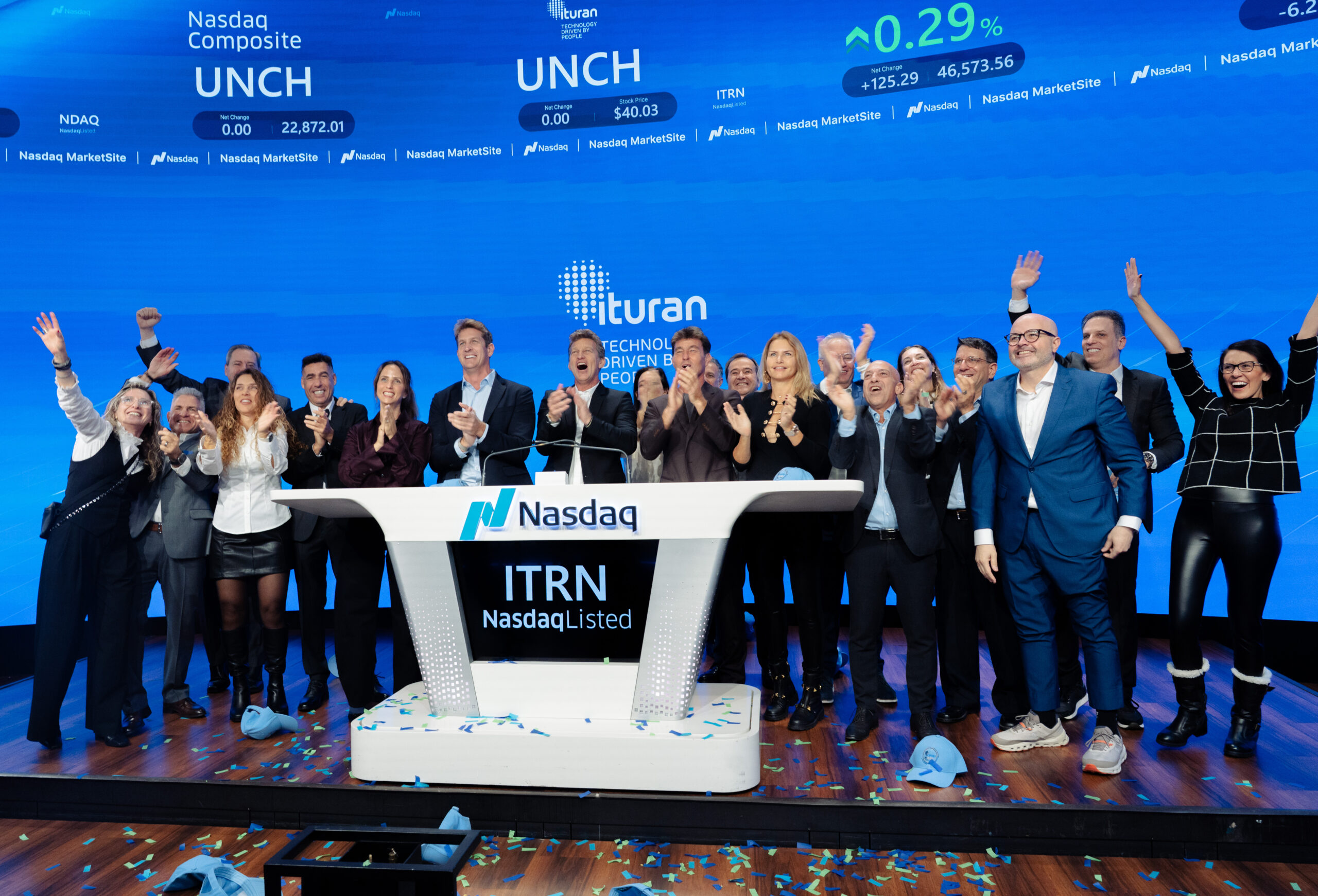 Equipo de Ituran celebrando en el escenario de Nasdaq tras tocar la campana de apertura, con pantallas azules y gráficos financieros de fondo