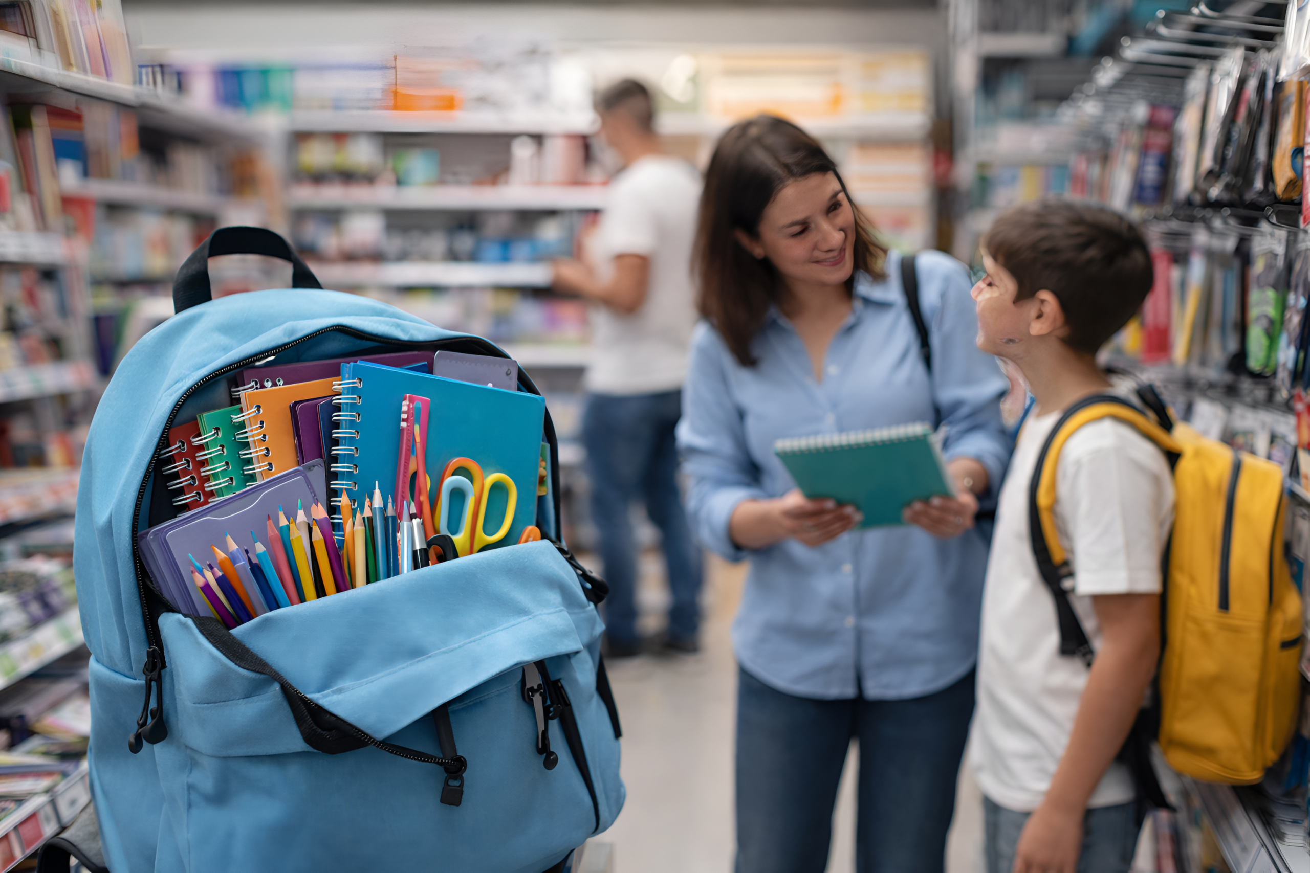 Mochila abierta con útiles escolares en primer plano mientras una madre y su hijo eligen productos en una librería