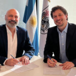 Dos representantes firmando un convenio institucional en una oficina con banderas argentinas de fondo
