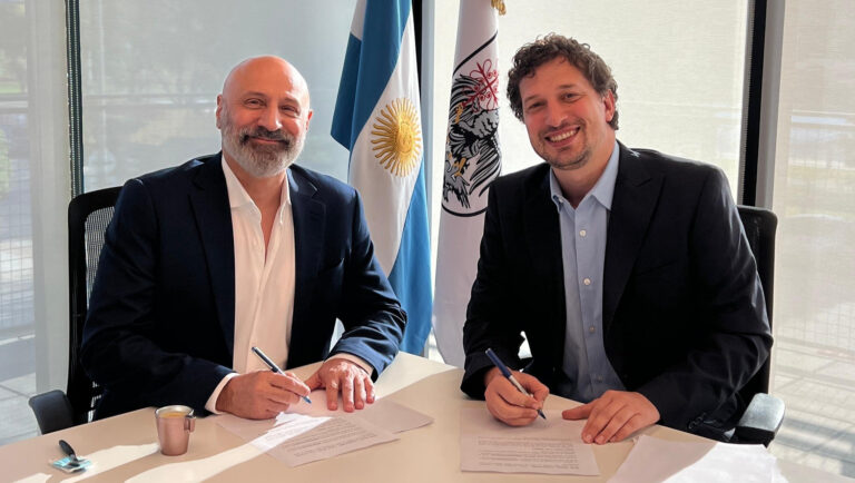Dos representantes firmando un convenio institucional en una oficina con banderas argentinas de fondo