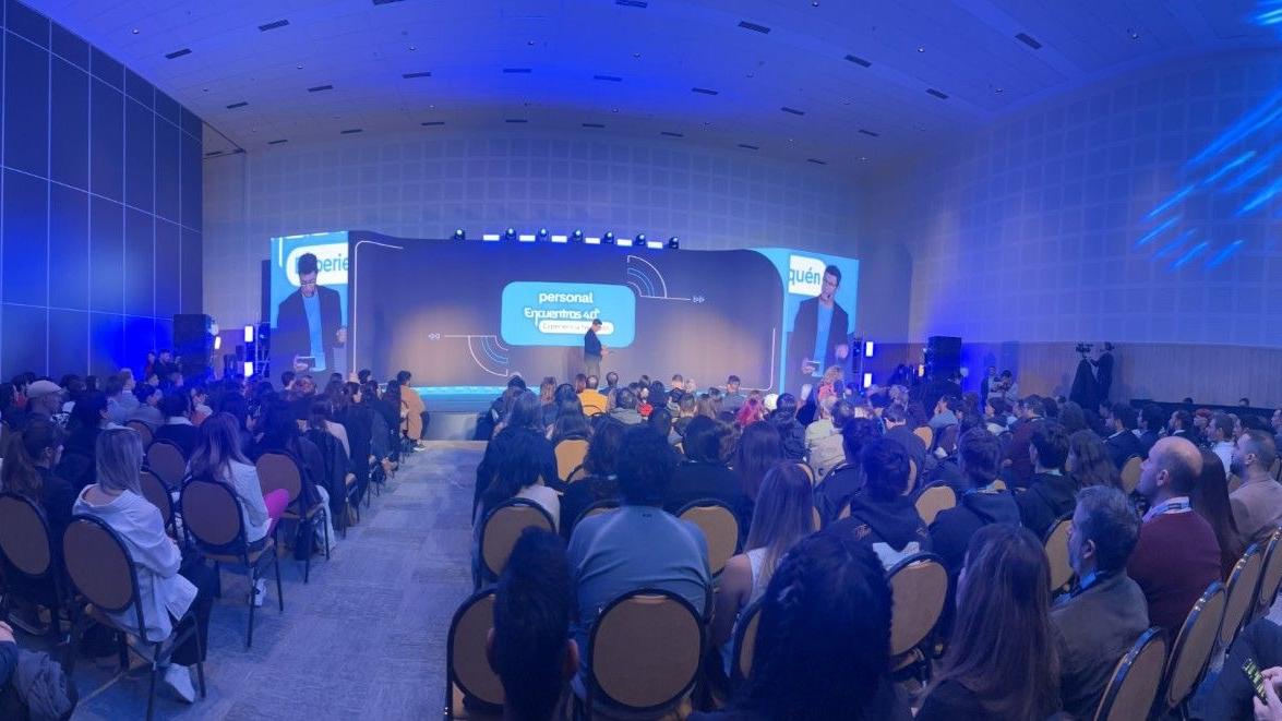 Auditorio lleno durante conferencia de tecnología con escenario iluminado y presentación sobre inteligencia artificial