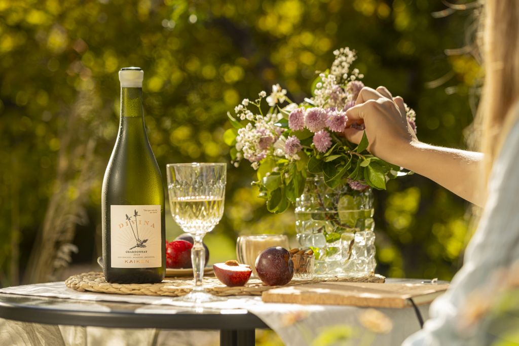 Botella de vino blanco Kaiken DIVINA junto a una copa y frutas en una mesa al aire libre rodeada de naturaleza y flores