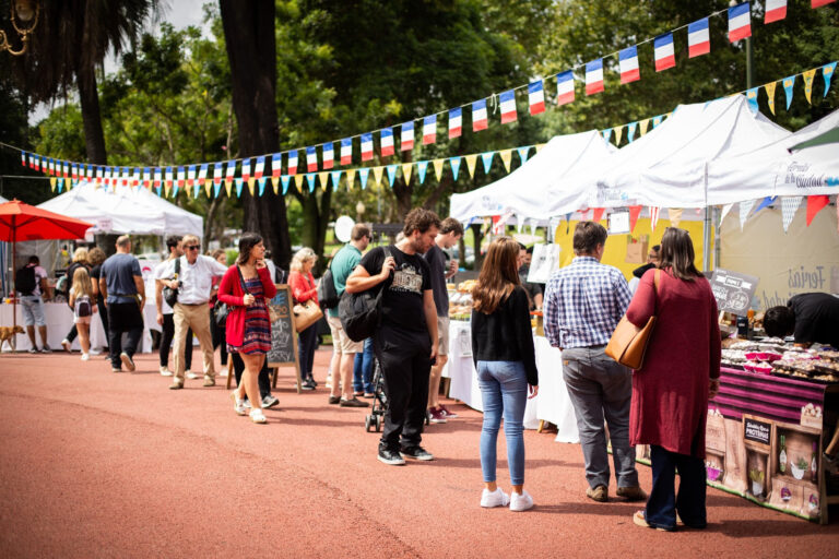 Personas recorriendo puestos gastronómicos en una feria al aire libre con banderas francesas en el Rosedal de Buenos Aires
