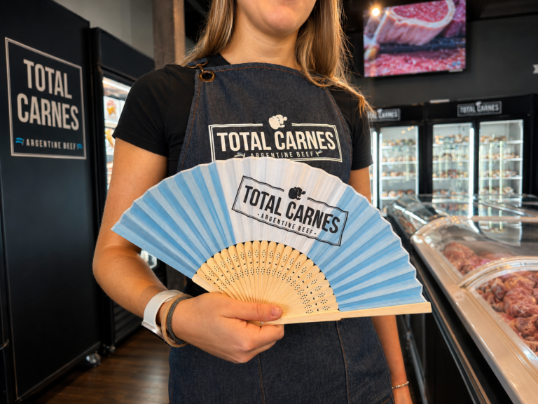Empleada de Total Carnes sostiene un abanico celeste y blanco con la marca de la empresa en una sucursal de Miami durante las activaciones especiales por el Mundial 2026