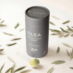 Envase de OLEA, la nueva infusión de Zuelo elaborada con hoja de olivo en polvo, presentada junto a hojas de olivo y una cucharilla con el producto