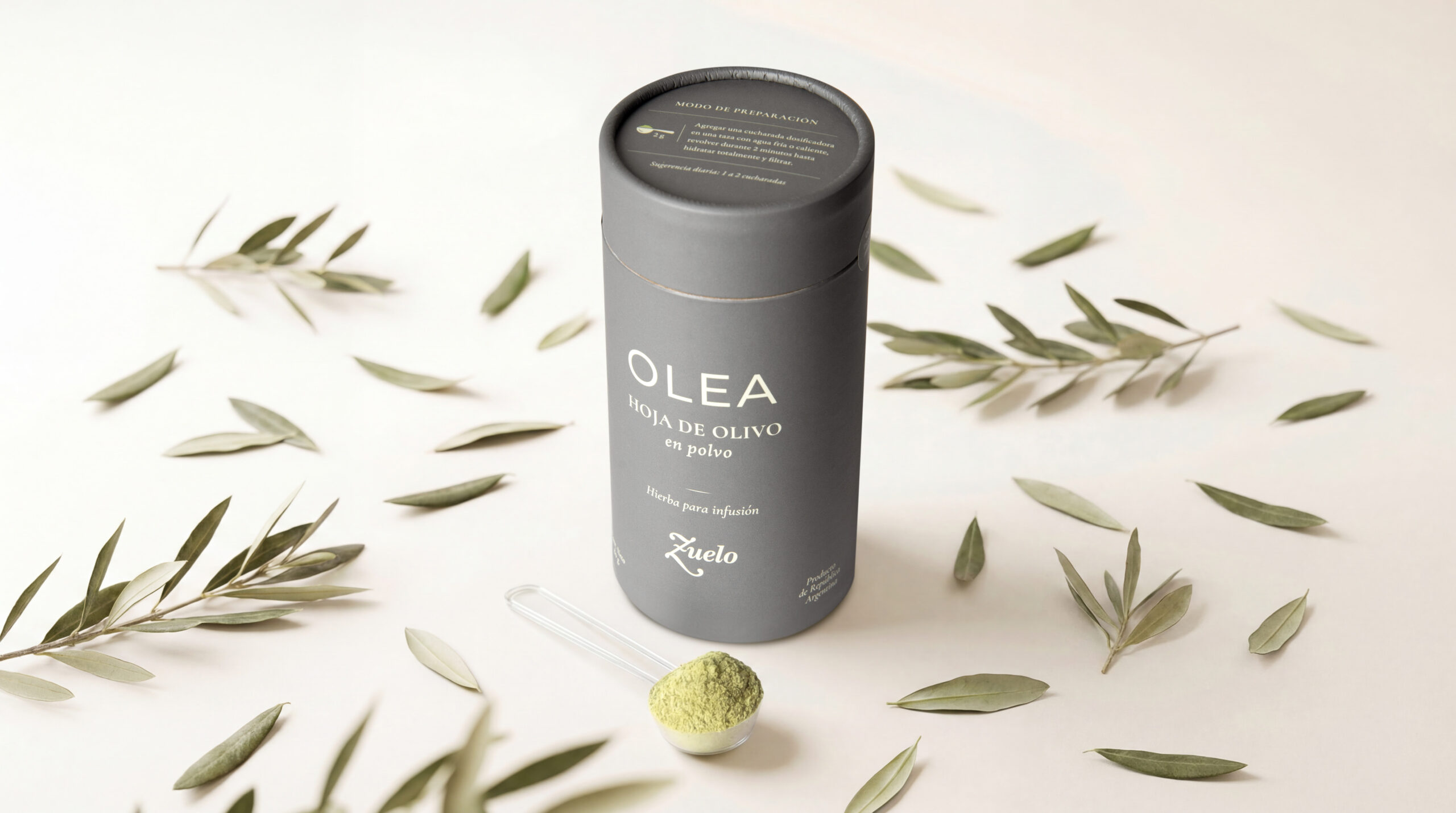 Envase de OLEA, la nueva infusión de Zuelo elaborada con hoja de olivo en polvo, presentada junto a hojas de olivo y una cucharilla con el producto