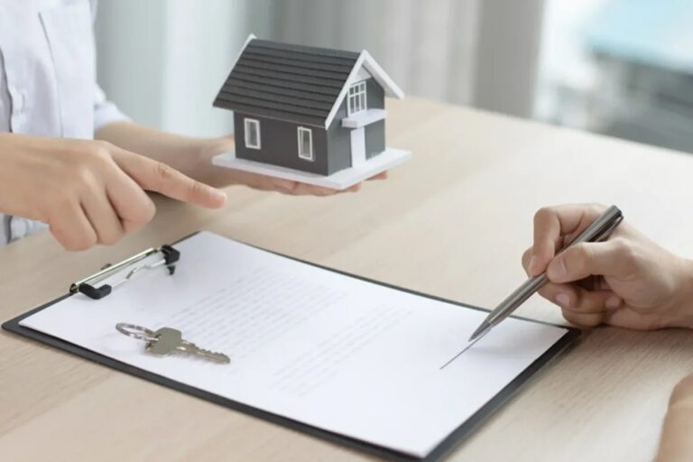 Persona firma la documentación para la compra de una vivienda mientras un asesor sostiene una maqueta de una casa y las llaves del inmueble sobre la mesa