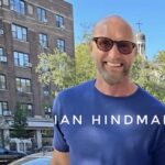 Ian Hindmarsh sonríe al aire libre durante el día, con edificios y árboles de fondo. Su nombre aparece sobreimpreso en la imagen