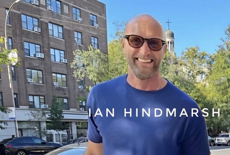 Ian Hindmarsh sonríe al aire libre durante el día, con edificios y árboles de fondo. Su nombre aparece sobreimpreso en la imagen