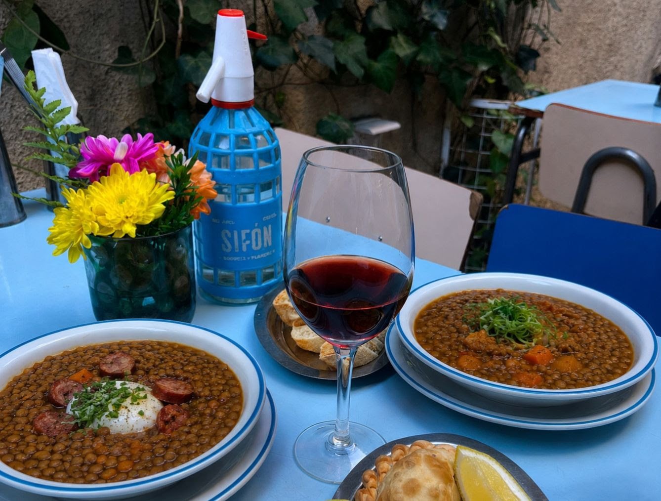 Guisos de lentejas servidos en platos junto a una botella de Sifón, pan y una copa de vino