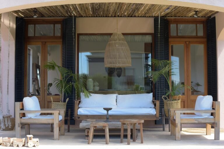 Living al aire libre con sofá y sillones de madera, almohadones blancos, plantas y lámpara colgante de fibras naturales, integrado a una vivienda con grandes ventanales