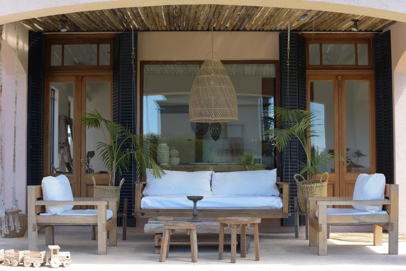 Living al aire libre con sofá y sillones de madera, almohadones blancos, plantas y lámpara colgante de fibras naturales, integrado a una vivienda con grandes ventanales